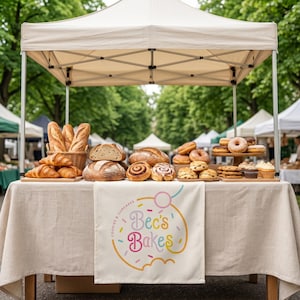 Op de afbeelding: Een marktkraam met een beige luifel met diverse bakproducten, waaronder baguettes in een mand, zuurdesembroden, kaneelbroodjes, donuts en cupcakes. Een witte banner aan de tafel bevat de tekst Bec's Bakes Cookies & Cupcakes.