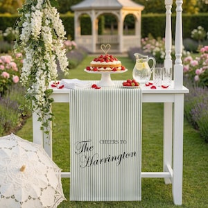 May include: A white table set for a garden party, featuring a cake with strawberries, a pitcher of lemonade, and glasses. A striped table runner reads "The Harrington". A white parasol and floral decorations complete the scene.