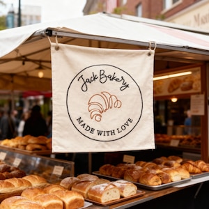 May include: A bakery stall banner with the text "Jack Bakery" in a circular design, featuring a croissant illustration and the words "Made with Love." Various breads and pastries are displayed on trays, ready for sale.