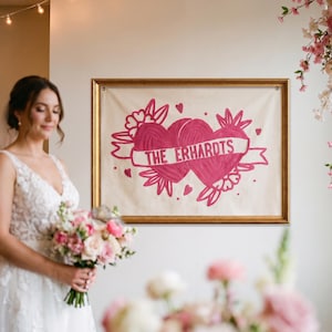 Op de afbeelding: Een rechthoekige wandhanger van gebroken witte stof in een gouden lijst, met een roze geborduurd ontwerp van twee verbonden harten met bloemaccenten en een banner in het midden met de tekst THE ERHARDTS in dikke roze hoofdletters.