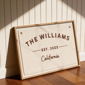 May include: A framed canvas sign with the text "THE WILLIAMS" above "EST. 2025" and "California" in a brown font. The canvas is beige, set in a light wood frame, and has brass grommets. The sign is leaning against a white wall.