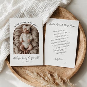 Pode incluir: Dois cartões retangulares brancos com texto preto e uma foto de um bebé a usar um body branco e uma fita com laço. Os cartões incluem o texto Auntie Amma & Uncle Mason, Will you be my Godparents?, um poema e o nome Baby Oliver.