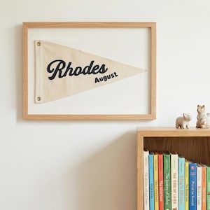 May include: A framed pennant flag, beige in colour, with the name "Rhodes" in black script and the month "August". The flag is mounted in a light wood frame. Decorative figurines and books are visible below the frame.