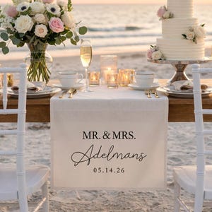 Puede incluir: Una mesa de recepción de boda en la playa, con un camino de mesa de lino blanco. El camino de mesa tiene el texto "MR. & MRS. Adelmans 05.14.26" en escritura negra. La mesa está decorada con porcelana blanca, cubiertos dorados, copas de champán y velas.