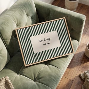 May include: A rectangular framed wall art piece featuring a light cream-colored center panel with the black text "how lucky are we" written in a cursive script. The border consists of vertical dark green and white stripes within a light wood frame.