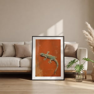 May include: A framed wall art print featuring a painting of a light blue and green gecko positioned diagonally against a solid, textured burnt orange background. The artwork is enclosed in a thin black frame with a white mat border.