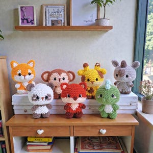 Könnte beinhalten: Eine Sammlung handgefertigter Häkeltier-Spielzeuge, darunter ein Fuchs, ein Affe, eine Giraffe, ein Nashorn, ein Panda und ein Dinosaurier. Die Spielzeuge sind in verschiedenen Farben wie Orange, Braun, Gelb und Grün gehalten und werden auf einem Holztisch präsentiert.