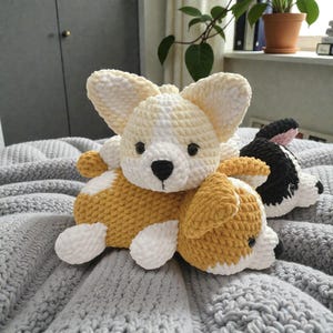May include: Crochet corgi dog plush toys in yellow, white, and black. The toys have large ears, black eyes, and a black nose. The corgis are lying on a grey knitted blanket. The plush toys are soft and cuddly.