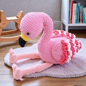 May include: A handmade pink crochet flamingo plush toy with a black beak, yellow accents, and a white and red striped tail. The flamingo is sitting on a gray rug.