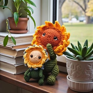 Puede incluir: Dos muñecos de girasol de ganchillo con caras sonrientes y tallos verdes están sentados en un alféizar de madera. El girasol más grande tiene pétalos naranjas y amarillos, mientras que el más pequeño tiene pétalos amarillos y azules. Una planta en maceta y libros están en el fondo.