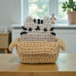 Może przedstawiać: Ręcznie robiona szydełkowa zabawka dla kota z czarnymi paskami i czarnymi guzikowymi oczami siedzi w beżowym szydełkowym koszu. Kot jest szary i biały. Kosz ma prostokątny kształt.