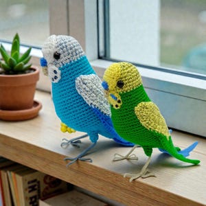 May include: Two handmade crochet parakeets. One is blue with white and gray accents, the other is green with yellow accents. Both have black eyes and are perched on a wooden surface. The birds are approximately 6 inches tall.
