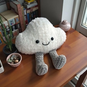 Könnte beinhalten: Ein flauschiges, weißes wolkenförmiges Kissen mit einem lächelnden Gesicht und grauen gehäkelten Beinen. Das Kissen besteht aus weichem Material und ist als Cartoon-Wolke gestaltet. Ein dekorativer Artikel für das Kinderzimmer.