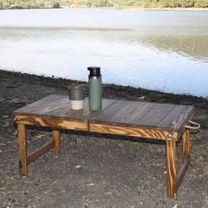 Può includere: Un tavolo da picnic pieghevole in legno con una macchia scura, allestito all'aperto vicino a un lago. Una bottiglia d'acqua verde e una tazza da viaggio grigia e bianca sono sul piano del tavolo. Il tavolo è lungo circa 76 cm.