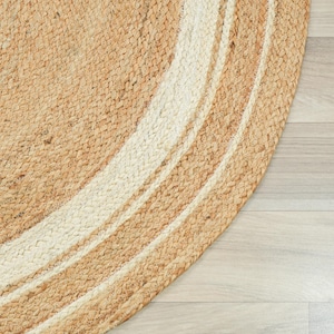 May include: A round braided rug featuring a natural tan jute texture with concentric circular bands of off-white fiber. The woven construction displays a thick, tactile pattern of interlaced fibers forming a circular border design.