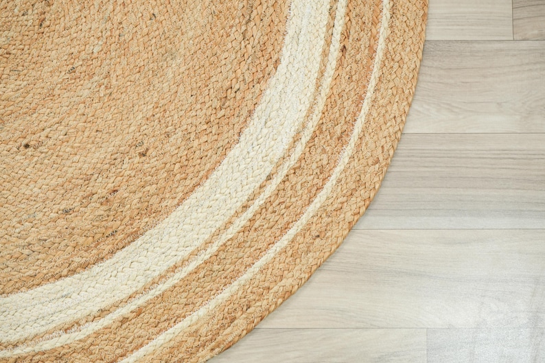 May include: A round braided rug featuring a natural tan jute texture with concentric circular bands of off-white fiber. The woven construction displays a thick, tactile pattern of interlaced fibers forming a circular border design.