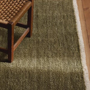 May include: A rectangular olive green rug with a textured, ribbed surface and white fringe along the edge, positioned next to a wooden stool featuring a woven seat. The rug displays a consistent, dense pile construction throughout its surface.