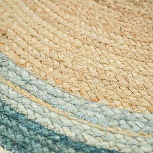 May include: A round woven rug featuring a natural tan jute center surrounded by concentric rings of light blue and dark teal fibers. The texture is created by thick, braided strands of plant-based material arranged in a circular pattern.