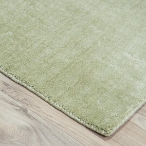 May include: A corner of a light green rug with a soft, textured surface. The rug's edge is visible, showing a neatly finished border. The rug is placed on a light-colored wooden floor, creating a contrast in textures and colors.