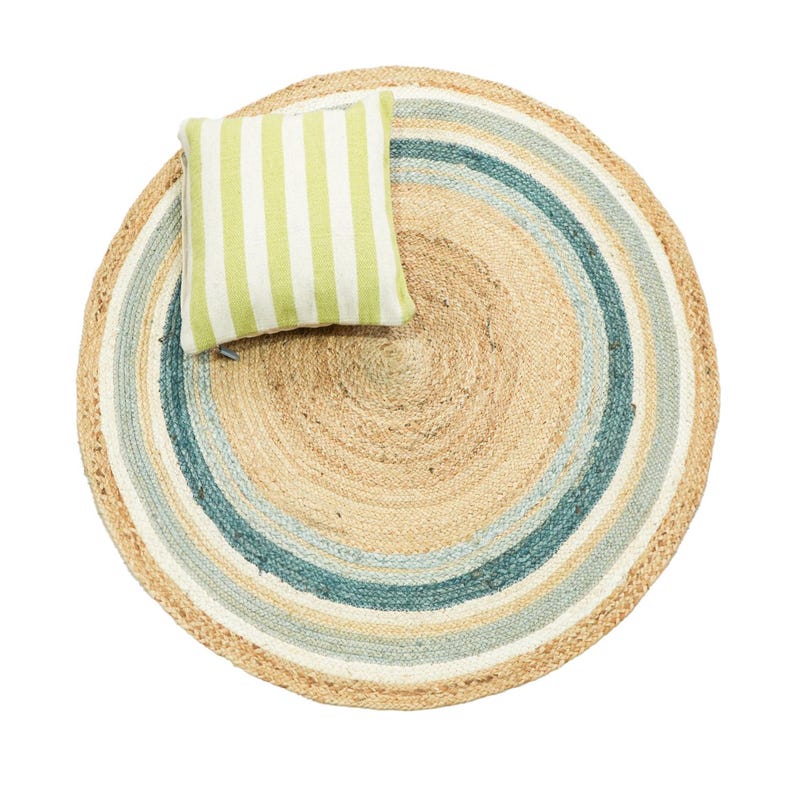 May include: A round, woven jute rug featuring concentric circles in natural tan, white, and teal blue. A square throw pillow with vertical lime green and white stripes is placed on top of the rug, creating a layered, textured visual composition.