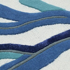 May include: A tufted rug featuring a white background with a raised, abstract pattern of organic, flowing shapes in shades of navy blue, royal blue, teal, and grey. The texture is dense and plush, with distinct color-blocked sections.