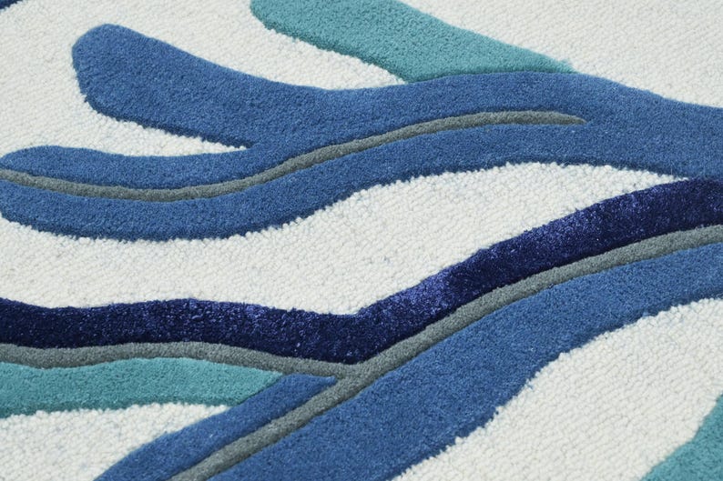 May include: A tufted rug featuring a white background with a raised, abstract pattern of organic, flowing shapes in shades of navy blue, royal blue, teal, and grey. The texture is dense and plush, with distinct color-blocked sections.