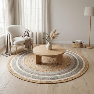 May include: A round, woven jute rug featuring concentric rings in natural tan, soft gray, and muted blue tones. The rug is placed on a light wood floor, serving as a base for a low, circular wooden coffee table with a smooth, flat surface.