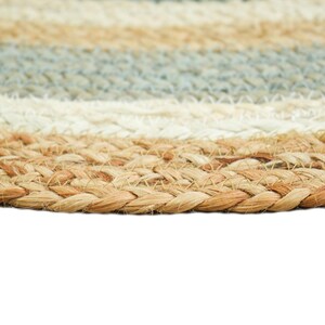 May include: A circular braided rug featuring concentric bands of natural tan, off-white, and muted blue fibers. The texture is composed of thick, woven plant-based strands arranged in a repeating pattern that creates a durable, layered surface.