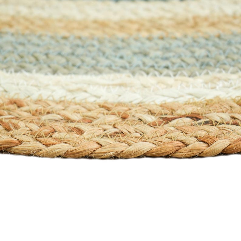 May include: A circular braided rug featuring concentric bands of natural tan, off-white, and muted blue fibers. The texture is composed of thick, woven plant-based strands arranged in a repeating pattern that creates a durable, layered surface.