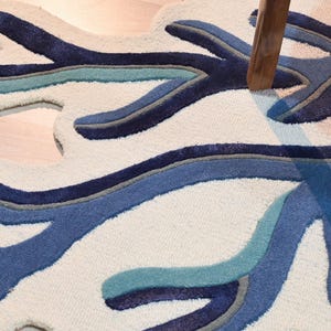 May include: A tufted area rug featuring an organic, branching coral-like shape. The design consists of an off-white background with layered, contoured patterns in shades of navy blue, medium blue, light teal, and grey, creating a textured surface.