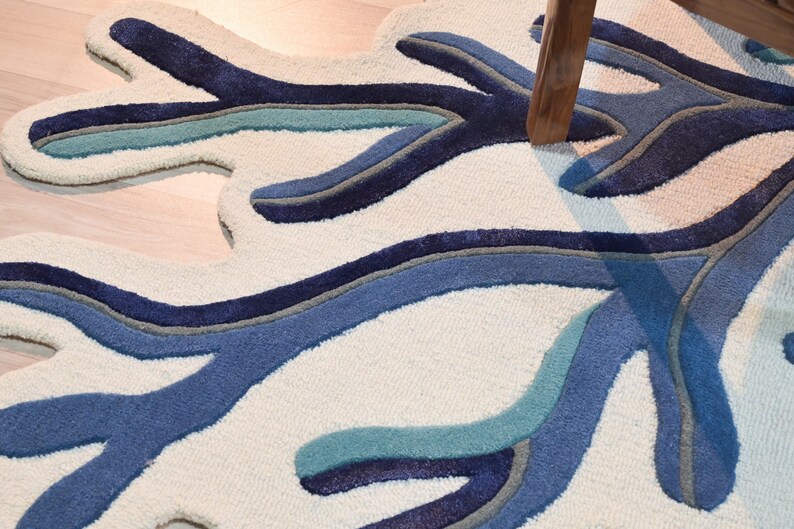 May include: A tufted area rug featuring an organic, branching coral-like shape. The design consists of an off-white background with layered, contoured patterns in shades of navy blue, medium blue, light teal, and grey, creating a textured surface.