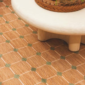 May include: A woven area rug featuring a tan base with a repeating grid pattern of white lines and small green squares at each intersection. The rug is positioned beneath a smooth, light-colored circular coffee table with a thick, rounded edge.