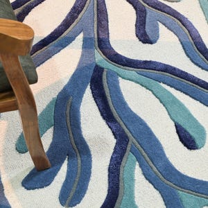 May include: A tufted area rug featuring a large-scale, branching coral-like pattern in shades of navy, royal blue, and teal against a pale cream background. The rug has a textured, cut-pile surface with defined edges between the colored sections.