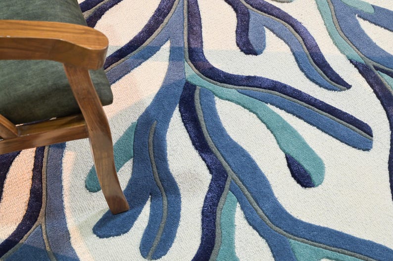 May include: A tufted area rug featuring a large-scale, branching coral-like pattern in shades of navy, royal blue, and teal against a pale cream background. The rug has a textured, cut-pile surface with defined edges between the colored sections.
