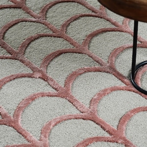 May include: A plush area rug featuring a repeating scalloped arch pattern in dusty rose pink against a light gray background, with a small portion of a wooden side table with black metal legs visible in the upper right corner of the frame.