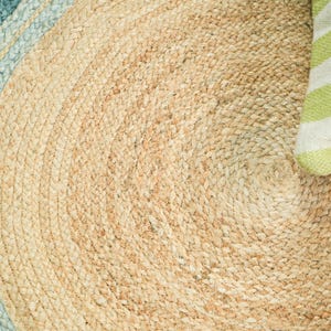 May include: A round braided rug made of natural tan fibers with a circular pattern. The outer edge features a wide band of light blue, while a small corner of the rug is partially covered by a white and lime green striped fabric element.