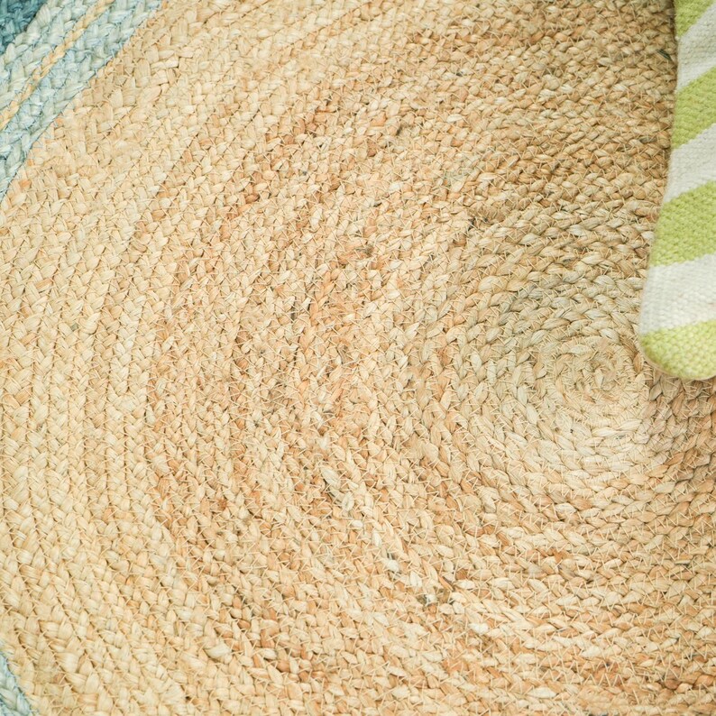 May include: A round braided rug made of natural tan fibers with a circular pattern. The outer edge features a wide band of light blue, while a small corner of the rug is partially covered by a white and lime green striped fabric element.