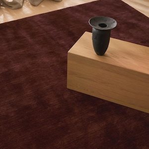 May include: A deep burgundy area rug with a subtle, uniform texture. A rectangular light-toned wooden block sits on the rug, supporting a dark, matte-finished vase with a wide, flared rim and a narrow, tapered base.