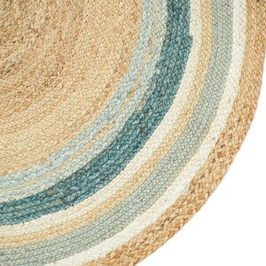 May include: A circular rug featuring a braided texture made from natural tan fibers. The design consists of concentric rings in alternating shades of light tan, soft white, and deep teal blue, creating a layered, circular pattern throughout the piece.