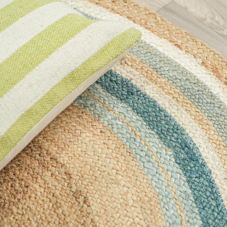 May include: A woven circular rug featuring concentric bands of natural tan, light blue, and cream fibers. A corner of a rectangular pillow with lime green and white diagonal stripes rests on top of the textured, braided surface of the rug.