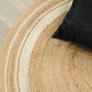 May include: A round, braided rug featuring concentric circles of natural tan and off-white fibers. A solid black, square-shaped pillow with a textured, fur-like surface rests on the right side of the rug, partially overlapping the circular pattern.