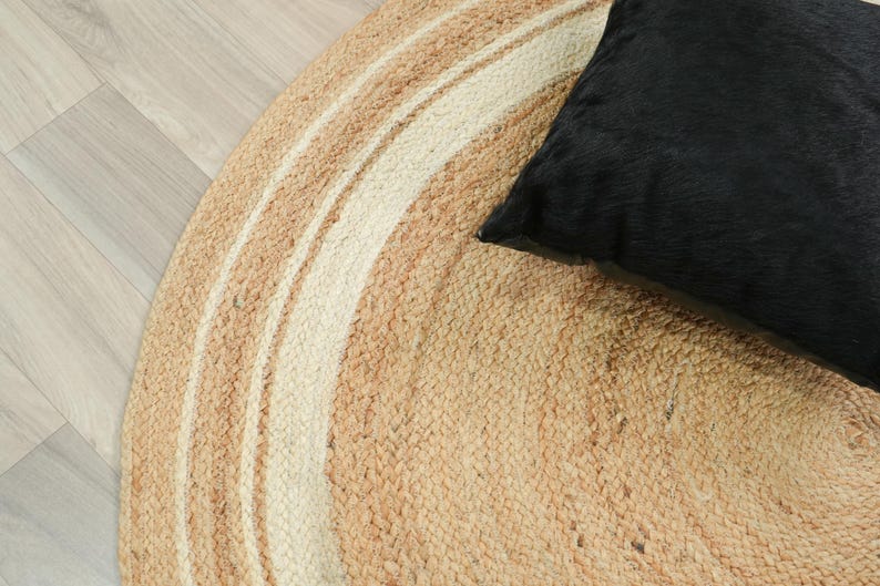 May include: A round, braided rug featuring concentric circles of natural tan and off-white fibers. A solid black, square-shaped pillow with a textured, fur-like surface rests on the right side of the rug, partially overlapping the circular pattern.