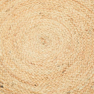 May include: A circular rug crafted from natural tan jute fibers, featuring a tightly woven braided pattern that spirals outward from the center. The texture is consistent and fibrous, showcasing the organic light brown tones of the plant material.