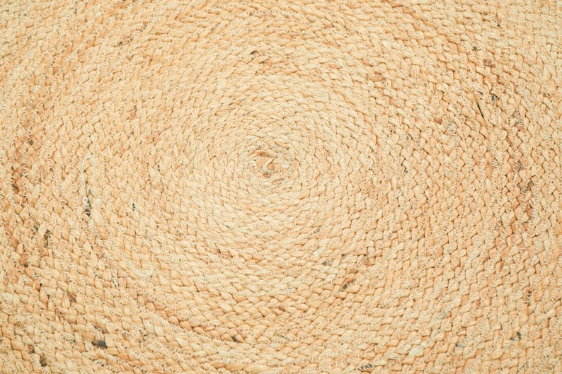 May include: A circular rug crafted from natural tan jute fibers, featuring a tightly woven braided pattern that spirals outward from the center. The texture is consistent and fibrous, showcasing the organic light brown tones of the plant material.
