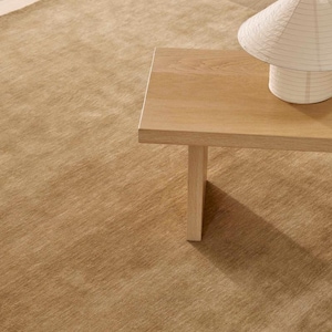 May include: A large, solid tan area rug with a short, dense pile texture. A light-colored wooden coffee table with a rectangular top and a single visible leg sits on the rug, supporting a white, conical paper lamp with a ribbed texture.