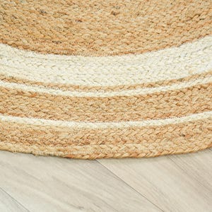 May include: A round area rug constructed from braided natural tan jute fibers and white cotton fibers. The design features concentric circular bands of tan and white, creating a textured, woven pattern that follows the curved edge of the rug.