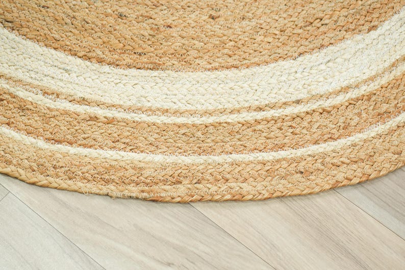 May include: A round area rug constructed from braided natural tan jute fibers and white cotton fibers. The design features concentric circular bands of tan and white, creating a textured, woven pattern that follows the curved edge of the rug.