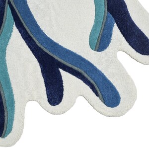 May include: A tufted rug with an irregular, organic shape featuring a cream-colored base. The surface is decorated with flowing, abstract lines in shades of navy blue, medium blue, and teal, creating a layered, wave-like pattern across the fabric.