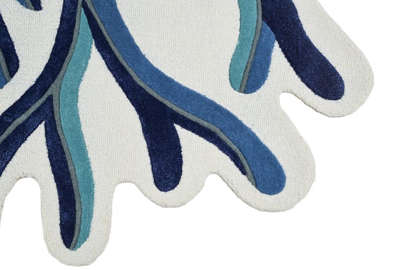 May include: A tufted rug with an irregular, organic shape featuring a cream-colored base. The surface is decorated with flowing, abstract lines in shades of navy blue, medium blue, and teal, creating a layered, wave-like pattern across the fabric.