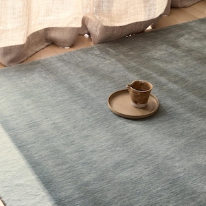 May include: A large, solid sage green area rug with a subtle, textured surface. Resting on the rug is a small, round, tan ceramic plate holding a single, small, brown, speckled ceramic pitcher with a spout and a slightly uneven, rustic finish.